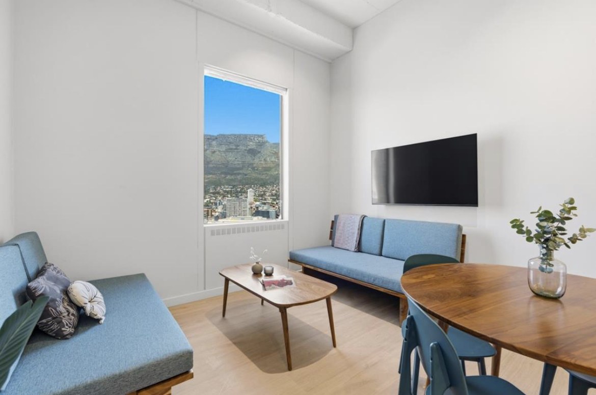 A bright, tidy apartment living room with blue sofas, a wooden table and a window view of Table Mountain